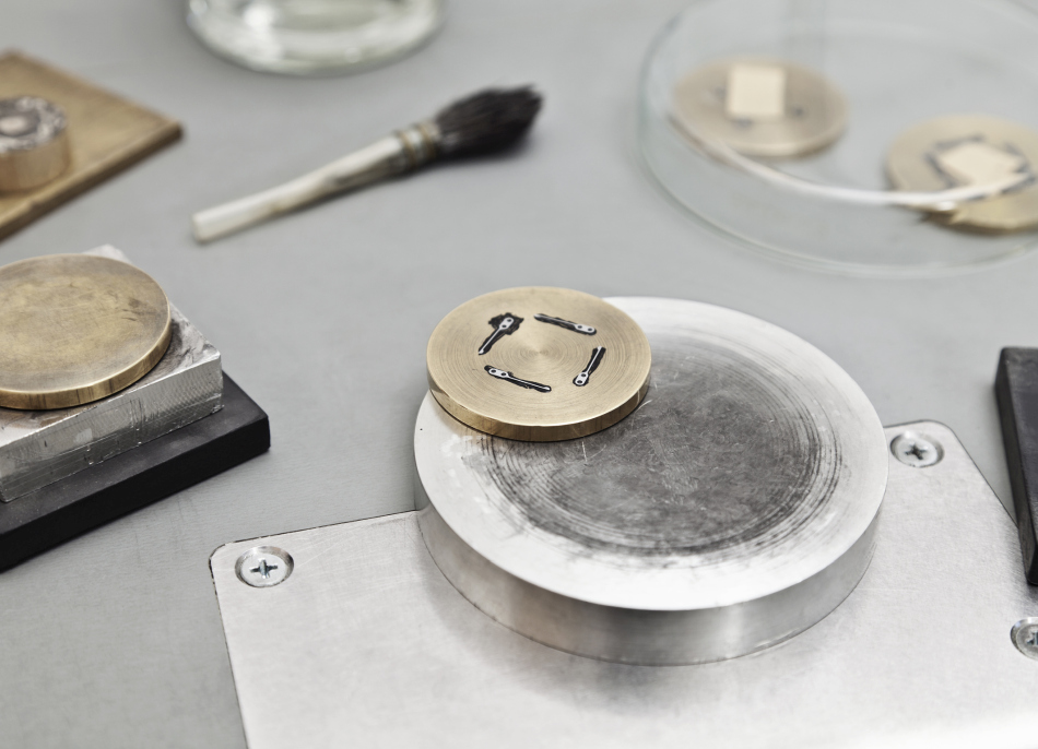 Polissage en noir : quatre ressorts de cliquet sont fixés à la cire d'abeille sur un anneau de laiton. Sur une plaque en étain, l'horloger polit la surface des ressorts avec de la pâte à polir jusqu'à ce que l'étain et la pâte deviennent noirs. Polissage en noir : quatre ressorts de cliquet sont fixés à la cire d'abeille sur un anneau de laiton. Sur une plaque en étain, l'horloger polit la surface des ressorts avec de la pâte à polir jusqu'à ce que l'étain et la pâte deviennent noirs.